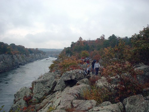 A rocky riverbank with people walking along the shore, surrounded by autumn foliage under a cloudy sky.