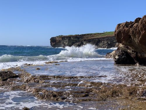 Waves crash against rocky shore under a clear blue sky, with a cliff in the background.