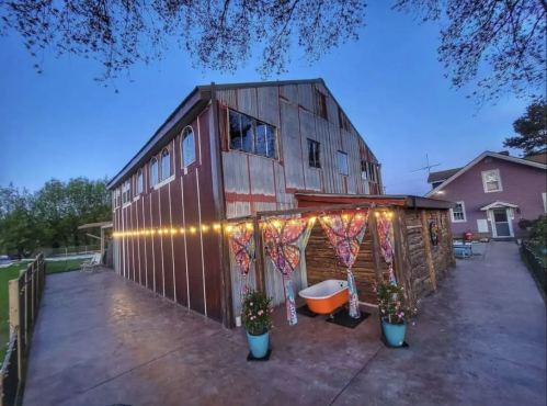 A rustic barn with colorful curtains, an orange bathtub, and string lights, set against a twilight sky.