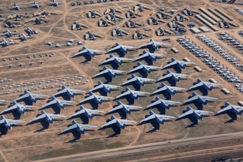 Aerial view of numerous military aircraft parked in a large, open area with additional structures in the background.