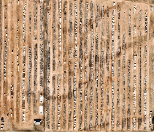 Aerial view of a large, organized junkyard filled with rows of vehicles and machinery on a dusty landscape.