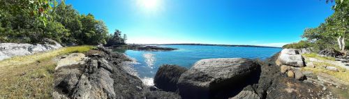 A panoramic view of a rocky shoreline with clear blue water and a bright sky, surrounded by lush greenery.