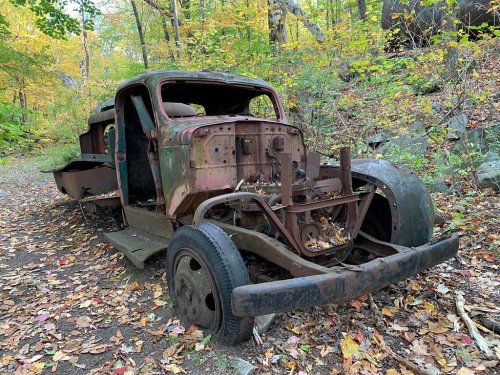 An old, rusted car chassis sits abandoned on a forest path, surrounded by colorful autumn leaves.