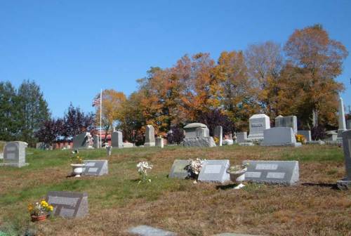 A peaceful cemetery with gravestones, colorful autumn trees, and an American flag in the background under a clear blue sky.