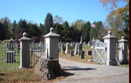 A cemetery entrance with stone pillars and a wooden stump, surrounded by gravestones and trees under a clear blue sky.