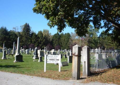 A view of Jewett City Cemetery, featuring gravestones and a "Please No Dogs" sign amidst trees and grass.