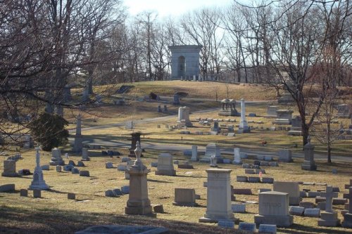 A serene cemetery landscape with various gravestones and monuments, surrounded by bare trees and a clear sky.