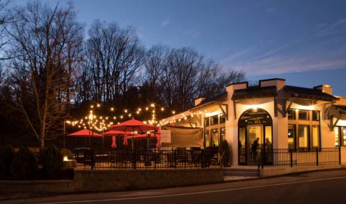 A cozy restaurant with outdoor seating, string lights, and red umbrellas, set against a twilight sky and bare trees.