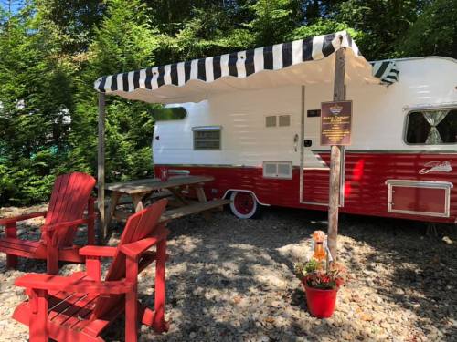 A vintage red and white camper with a striped awning, surrounded by greenery and featuring red Adirondack chairs.