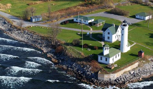 Aerial view of a lighthouse and surrounding buildings by the shore, with waves crashing against the rocky coastline.