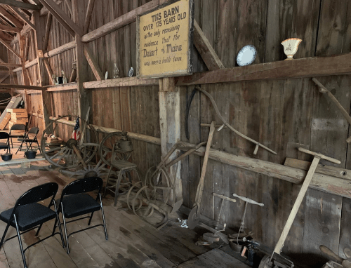 Interior of a rustic barn with old farming tools, chairs, and a sign about the barn's history on the wall.
