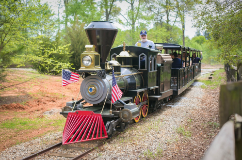 A vintage steam train with a passenger car, decorated with American flags, traveling along a gravel track in a green landscape.
