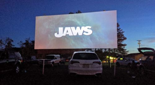 A drive-in movie screen displaying the title "JAWS" at dusk, with cars parked in front.
