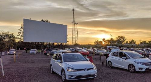 A drive-in movie theater at sunset, with cars parked and a large blank screen in the background.