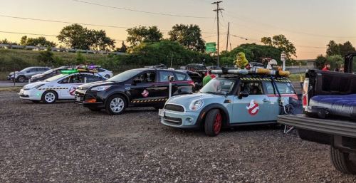 Three themed cars parked in a gravel lot, featuring Ghostbusters decorations and colorful lights, with a sunset backdrop.