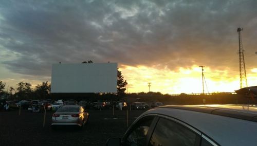 A drive-in movie theater at sunset, with cars parked and a large blank screen in the foreground.