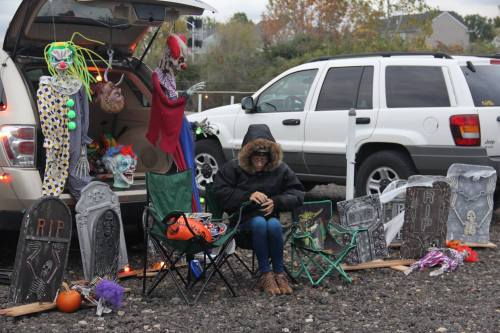 A person in a hooded jacket sits in front of a car, surrounded by Halloween decorations, including tombstones and clowns.