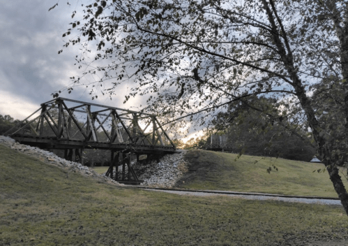A rustic metal bridge spans a grassy area, framed by trees and a cloudy sky at sunset.