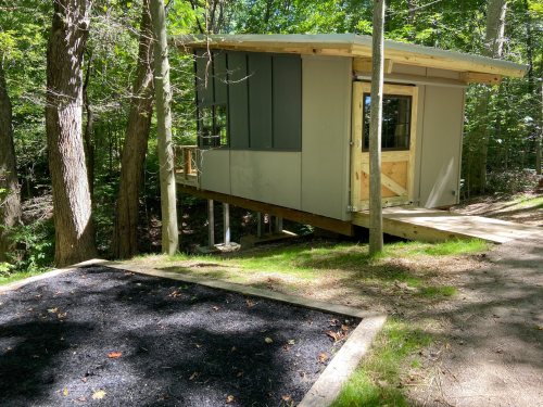 A modern cabin on stilts in a wooded area, with a path leading to it and a black gravel area in front.