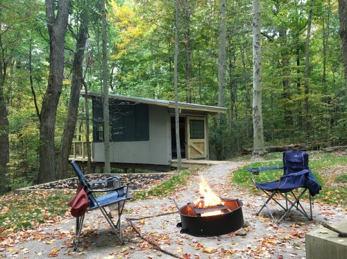 A cozy cabin in the woods with a fire pit and two camping chairs surrounded by autumn foliage.