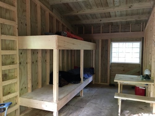 Interior of a wooden cabin with bunk beds, a table, and a window, featuring unfinished wooden walls and floor.