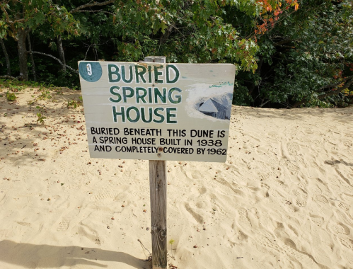 Sign reading "Buried Spring House" with information about a spring house built in 1938, now covered by sand.