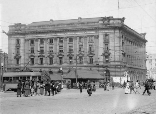 Historic black-and-white photo of a grand building with people and a streetcar in front, bustling urban scene.