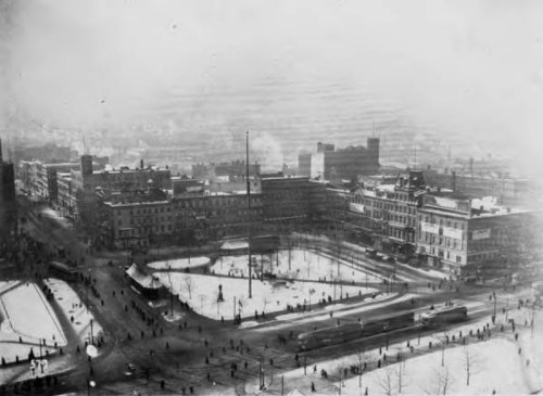 Aerial view of a snowy city square with people walking, buildings, and a flagpole in the center. Black and white photo.