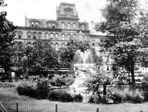 Historic black-and-white photo of a park with a fountain, surrounded by trees and a building in the background.