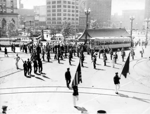 A historic parade with marching bands and flag bearers in a city square, featuring a streetcar in the background.