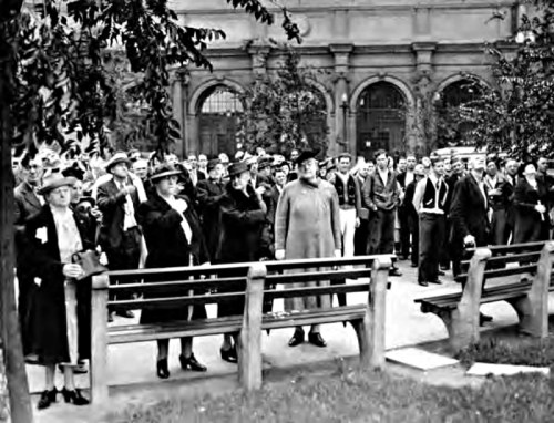 A crowd of people in early 20th-century attire gathered in a park, some looking up, with benches in the foreground.