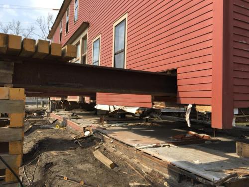 A red house elevated on supports, revealing a steel beam and construction materials underneath.