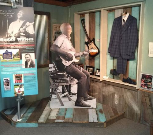 A life-size cutout of a guitarist in a museum exhibit, featuring a suit and guitar on display.