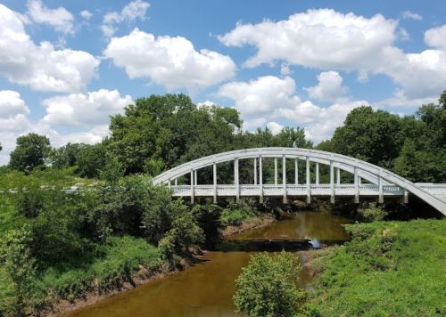 A white arch bridge spans a calm river, surrounded by lush greenery and a bright blue sky with fluffy clouds.