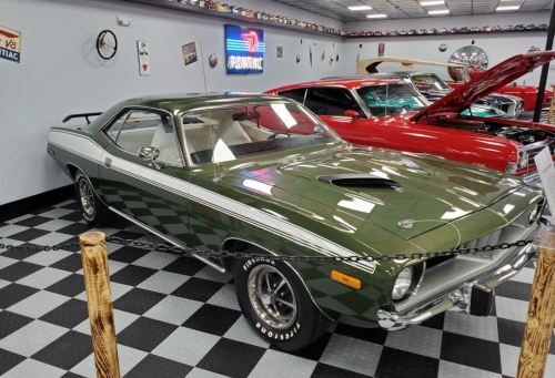 A classic green muscle car displayed in a showroom with checkered flooring and other vintage cars in the background.