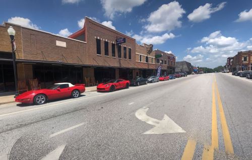 A street view featuring several red sports cars parked along a brick building with a blue sky and fluffy clouds.