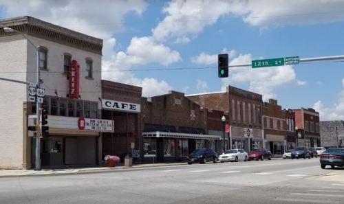 A small town street featuring historic buildings, a cafe, and a movie theater under a blue sky with fluffy clouds.