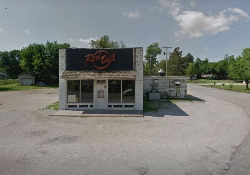 A small, single-story building with a sign reading "Red Ball," surrounded by gravel and trees on a sunny day.