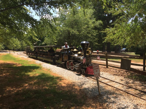 A vintage steam train with a passenger car travels along a gravel track surrounded by trees and a wooden fence.