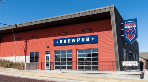 Exterior of a brewpub building with a red facade, large "BREWPUB" sign, and an "OPEN" banner. Clear blue sky above.
