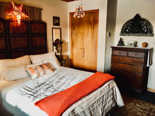 Cozy bedroom with a white bed, orange throw, wooden dresser, and decorative accents, creating a warm atmosphere.