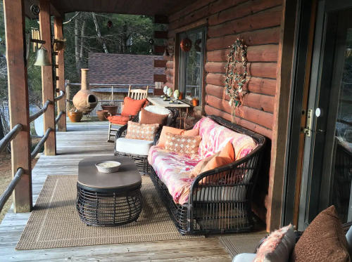 Cozy porch with a wicker sofa, coffee table, and rustic decor, surrounded by trees and a log cabin.