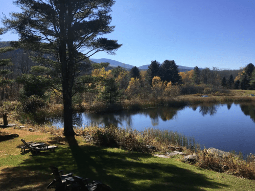 A serene lake surrounded by trees and mountains, with clear blue skies and autumn foliage reflecting on the water.
