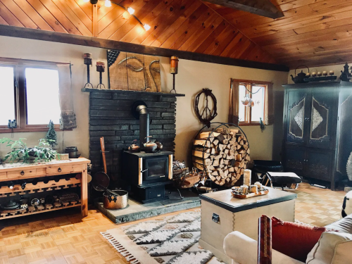 Cozy living room with a wood stove, stacked firewood, wooden furniture, and rustic decor under a wooden ceiling.