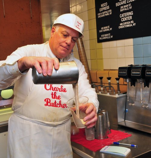 A man in a white apron and hat pours a milkshake into a cup at a diner counter.