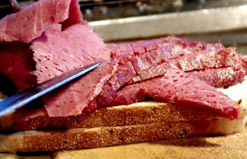 Close-up of sliced corned beef on bread, with a knife resting beside it, on a wooden cutting board.
