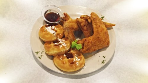A plate featuring crispy fried chicken, waffles, and a small cup of syrup, garnished with powdered sugar and parsley.