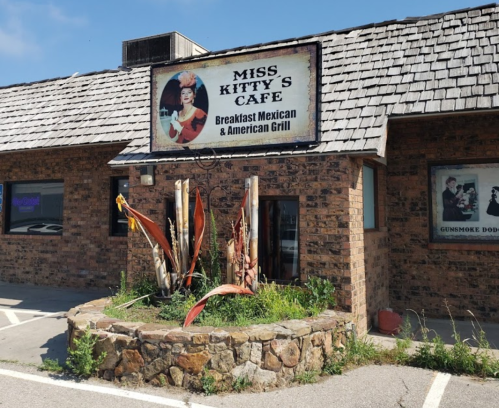 Sign for Miss Kitty's Cafe, featuring a woman in a hat, with a stone planter and colorful plants in front.