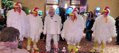 A group of people in chicken costumes stands in a casino, with a man in a white suit and glasses in the center.