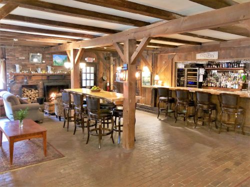 Cozy rustic bar area with wooden beams, a fireplace, and a long counter with stools in a warmly lit setting.